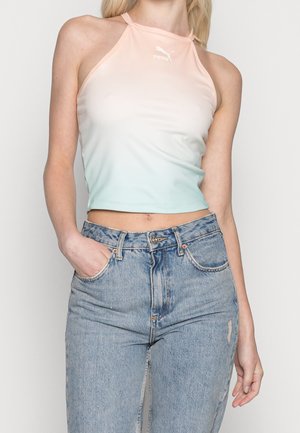 Frau trägt ärmelloses pastellfarbenes Ombre-Puma-Crop-Top mit Stehkragen und hellblauen hoch taillierten Jeans, Hand in der Tasche, steht vor weißem Hintergrund.