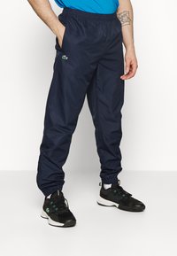 Marineblå nylon joggebukser med elastisk midje og mansjetter, med sidelommer og en liten grønn logo på venstre lår.