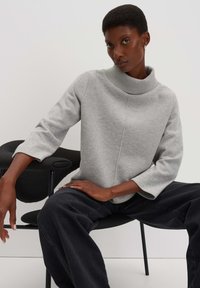 Grauer Sweatshirt mit hohem, geripptem Kragen und kurz geschnittenen Ärmeln, das eine glatte Textur und ein minimalistisches Design aufweist. Über dunklen Hosen getragen.