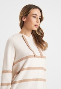 Hoodie w kolorze kremowym, wykonany z miękkiego dzianinowego materiału, z poziomymi beżowymi paskami, kapturem ze sznurkiem oraz luźnym krojem.