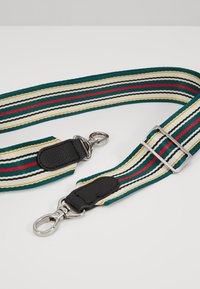 Tkani textilní pásek v pruzích zelené, krémové, červené a černé barvy, s kovovými stříbrnými detaily a koženými doplňky na upevňovacích místech.