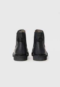 Bottines en cuir noir avec une finition lisse, des orteils arrondis et des talons empilés. À l'arrière, des languettes avec un détail de logo en relief.