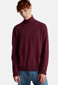 Burgundowy sweter z golfem charakteryzuje się miękką plecionką, długimi rękawami i dopasowanym krojem, zestawiony z jasnoniebieskimi dżinsami.