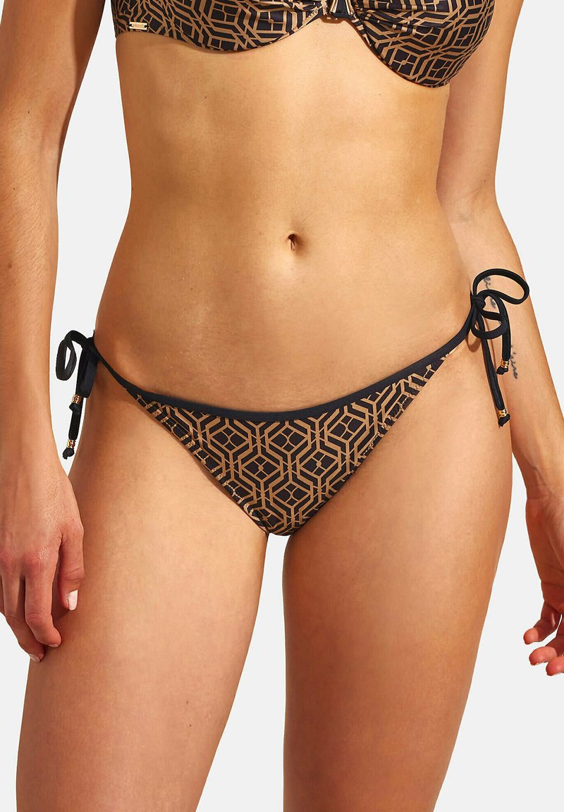 Vrouw in geometrisch patroon zwart en tan bikini onderkant, vastgebonden met strikken aan beide zijden, die de buik en handen toont.