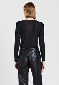 Schwarze Bluse mit langen Ärmeln und einem strukturierten gepunkteten Muster, aus leichtem Stoff und mit lockerer Passform, getragen zu glänzenden schwarzen Hosen.