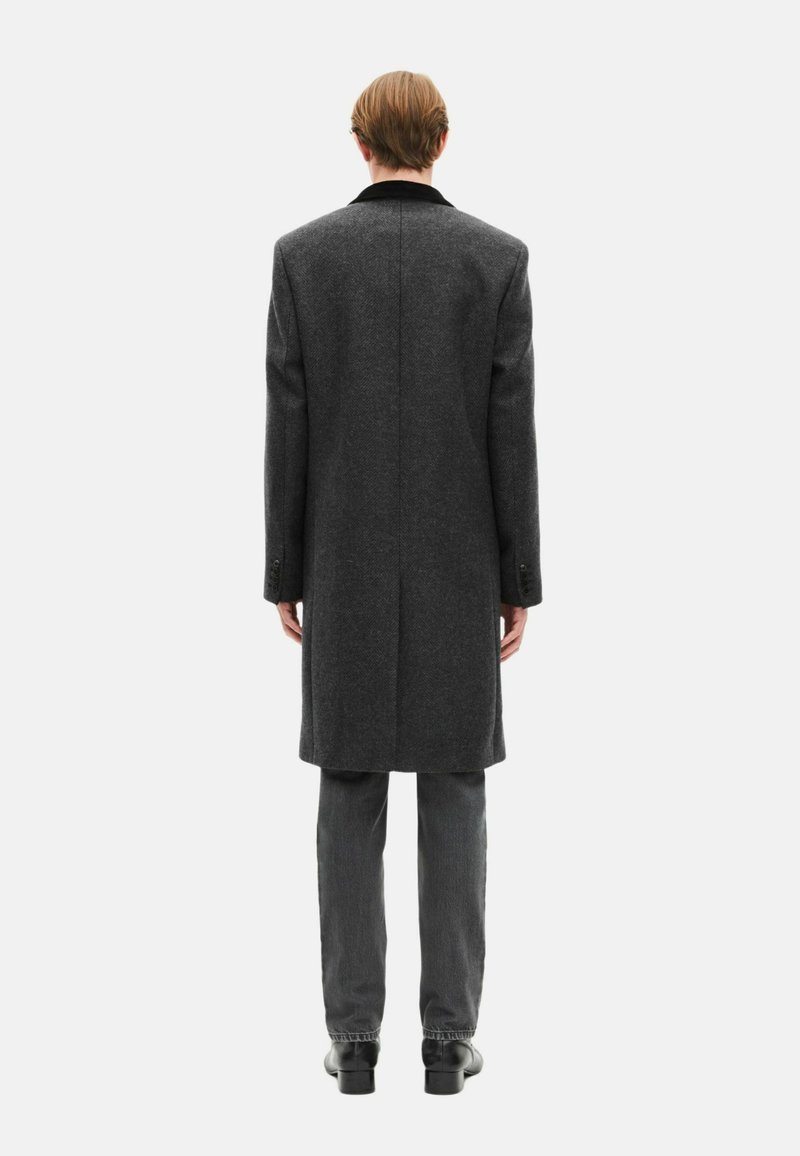 Long gray wool coat with a notched collar, single vent, and two-button cuffs, paired with dark gray trousers and black shoes.