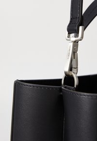 Geantă de mână din piele neagră cu o textură netedă, având o curea robustă și accesorii de nuanță argintie. Cusături detaliate vizibile pe margini.