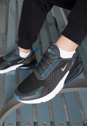 Person iført sorte og blå Nike Air Max 270 sneakers og hvide sokker, siddende på en metalbænk med krydsede ben.