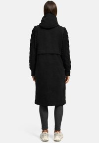 Black long coat featuring a hood, braided texture on sleeves, and a sleek design. Worn over dark leggings and light sneakers.