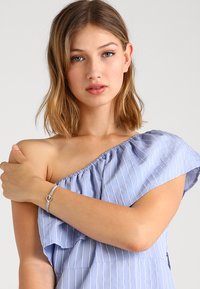 Blau gestreiftes Off-Shoulder-Top mit Rüschendetails. Das Model trägt ein silbernes Armband mit einem kreisförmigen Akzent an ihrem Handgelenk. Glatter Stofftextur.
