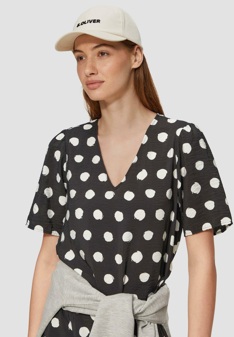 Zwarte shirt met witte stippen, V-hals ontwerp, korte mouwen en een textuurstof. Het model draagt een beige pet met zwarte letters.