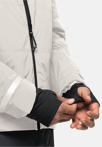 Des mains ajustant des poignets de manches intérieures noirs sous une veste gris clair avec fermeture éclair sur le devant et bande réfléchissante sur la manche.