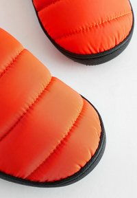 Orange quiltad toffla med en slät, vadderad ovandel och en texturerad svart gummisula. Har sydda detaljer och rundad tådesign.