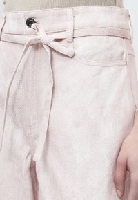 Ανοιχτό ροζ παντελόνι denim με υφασμάτινη ζώνη δεμένη σε φιόγκο στη μέση, φορεμένο με μπεζ τοπ και ορατό χέρι στο πλάι.