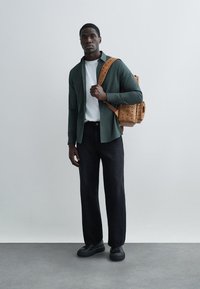 Young man wearing a green open shirt over white tee, black pants, black shoes, carrying a tan studded backpack on one shoulder.