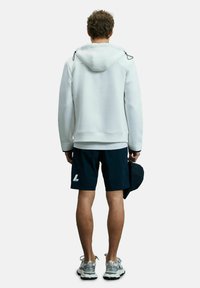 Weiße Kapuzenjacke mit Kordeln, kombiniert mit dunkelblauen Shorts mit einem Logo. Das Modell hält eine schwarze Mütze, während silberne Laufschuhe sichtbar sind.