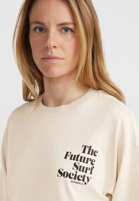 Beige bavlněné tričko s volným střihem, na levé části hrudi má výrazný černý grafický text "The Future Surf Society O'Neill."