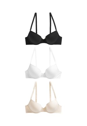 Młoda kobieta z długimi włosami, ubrana w beżowy biustonosz z wkładkami, patrząca lekko na bok, na jednolitym jasnym tle.