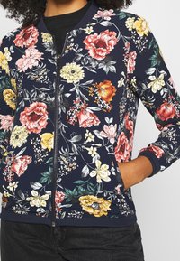 Veste bomber florale avec un fond bleu marine, ornée de fleurs roses, jaunes et rouges. Poignets côtelés et détail de fermeture éclair à l'avant.