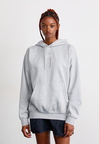 Jeune femme avec des cheveux tressés portant un sweat à capuche gris clair et une jupe noire, se tenant devant un fond blanc uni.