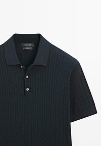 Tmavě modrá bavlněná polokošile s texturovaným vzorem copánkového pletení, krátkými rukávy a tříknopovým rozparkem. Na límci je viditelná štítek.