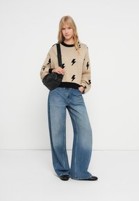 Béžový sveter s čiernymi vzormi bleskov, v kombinácii s voľnými modrými džínsami, doplnený o čiernu crossbody tašku a čierne topánky.