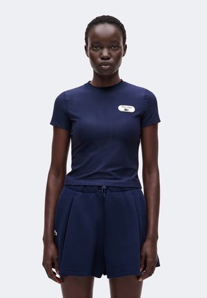 Vrouw draagt een getailleerd navy ribgebreid kortarmig topje en losse navy shorts met een koord in de taille, staand tegen een effen achtergrond.
