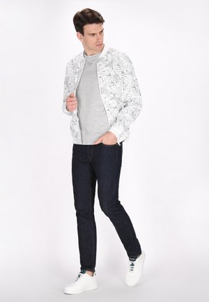 Chaqueta bomber ligera con fondo blanco y patrón de hojas negras, combinada con una camiseta de manga larga gris, jeans oscuros y zapatillas blancas.