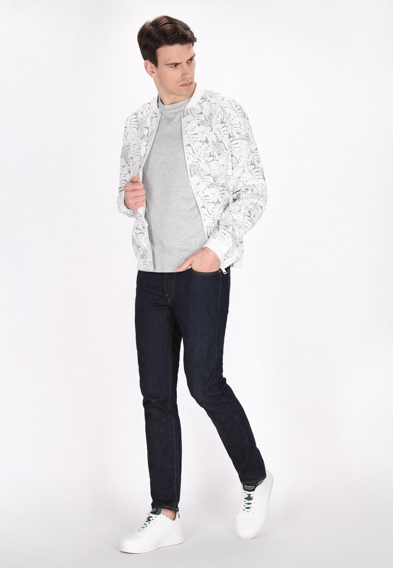 Lichte bomberjack met een witte achtergrond en zwart bladeren patroon, gecombineerd met een grijs longsleeve shirt, donker jeans en witte sneakers.