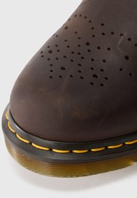 Καφέ δερμάτινο παπούτσι με άκρη με τρυπητό μοτίβο, κίτρινη ραφή και διάφανη καουτσούκ σόλα σε λευκό φόντο.