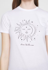 Camiseta blanca de algodón que presenta un dibujo lineal negro del sol con olas y estrellas, con la leyenda "brilla como el sol" debajo de la imagen.