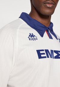 Baltas sportinis marškinėlis su tamsiai mėlynomis apykaklėmis. Su mėlynu Kappa logotipu ir ryškiais tamsiai mėlynos spalvos užrašais „EMΣ“. Švelnios tekstūros, kasdienio stiliaus.