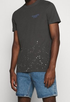 Ciemnoszara bawełniana koszulka z białym wzorem rozprysków na dole, z logo "Abercrombie & Fitch" w kolorze niebieskim, zestawiona z dżinsowymi szortami.