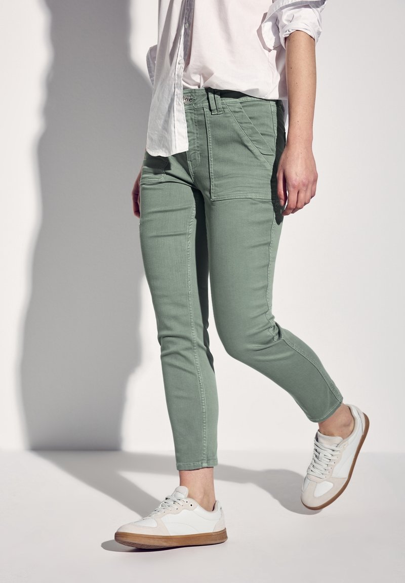 Personne portant une chemise blanche retroussée, un pantalon slim vert sauge et des baskets blanches avec semelles marron clair, marchant devant un fond uni.