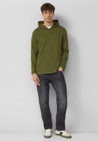 Olijfgroene hoodie met een voorzak, lange mouwen en een hoge kraag, gecombineerd met donkergrijze spijkerbroek en witte sneakers met accenten.