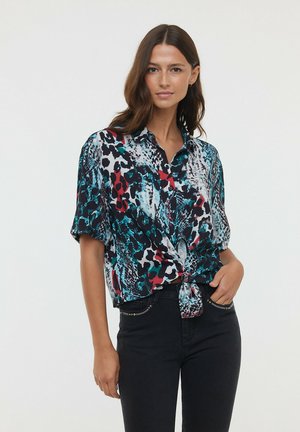 Blouse à manches courtes avec un nœud sur le devant, arborant un imprimé animalier abstrait coloré dans des tons de noir, rouge, turquoise et blanc.