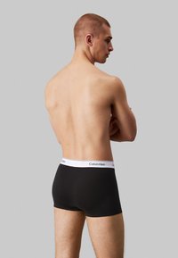 Svart boxer-truser med en hvit elastisk midjebånd som har "Calvin Klein"-merke, myk stoff, tettsittende fasong og lengde til midt på låret.