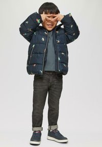 Marine pufferjack met dinosaurustpatronen, grijze hoodie, donkere jeans en blauwe sneakers. Kind poseert met vingers die een driehoek vormen.