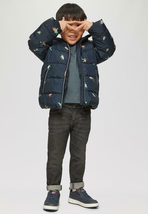 Marineblauer Pufferjacke mit Dinosauriermustern, grauem Hoodie, dunklen Jeans und blauen Sneakern. Kind posiert mit Fingern, die ein Dreieck bilden.
