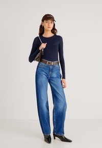 Top de manga larga azul marino, cinturón marrón, vaqueros azules de talle alto y sueltos, botines negros puntiagudos y gorra marrón. Bolso texturizado con cadena.