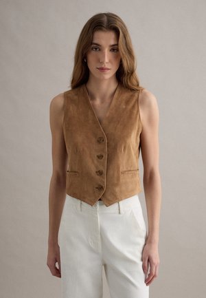 Donna con lunghi capelli castani, che indossa un gilet smanicato in camoscio beige con bottoni e pantaloni bianchi a vita alta, in piedi su uno sfondo neutro.