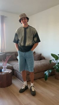 Νεαρός άνδρας φοράει μπλουζάκι tie-dye, τζιν σορτς, μπεζ καπέλο, λευκές κάλτσες και μαύρα αθλητικά παπούτσια, στέκεται μέσα σε εσωτερικό χώρο κοντά σε έναν καναπέ και φυτά.
