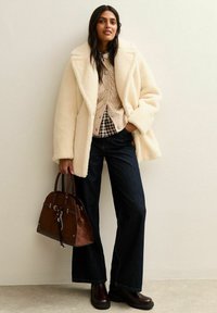 Cappotto in pelliccia sintetica color crema, maglione beige a trecce, camicia a quadri, jeans a gamba larga blu scuro, borsa marrone e stivaletti marroni.