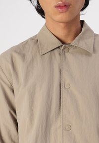 Chaqueta ligera beige con cuello, que presenta cierres a presión y un tejido texturizado. El diseño se centra en la simplicidad y funcionalidad.