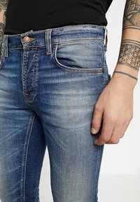 Mörkblå denimjeans med lätt slitningar, orange sömmar, fem fickor och en slim fit-stil. Inkluderar robust metallknappstängning.