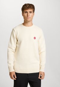 Kremgenser med teksturert stoff, ribbestrikkede mansjetter og kant, med en liten rød sirkulær logo på brystet. Brukt sammen med svarte bukser.