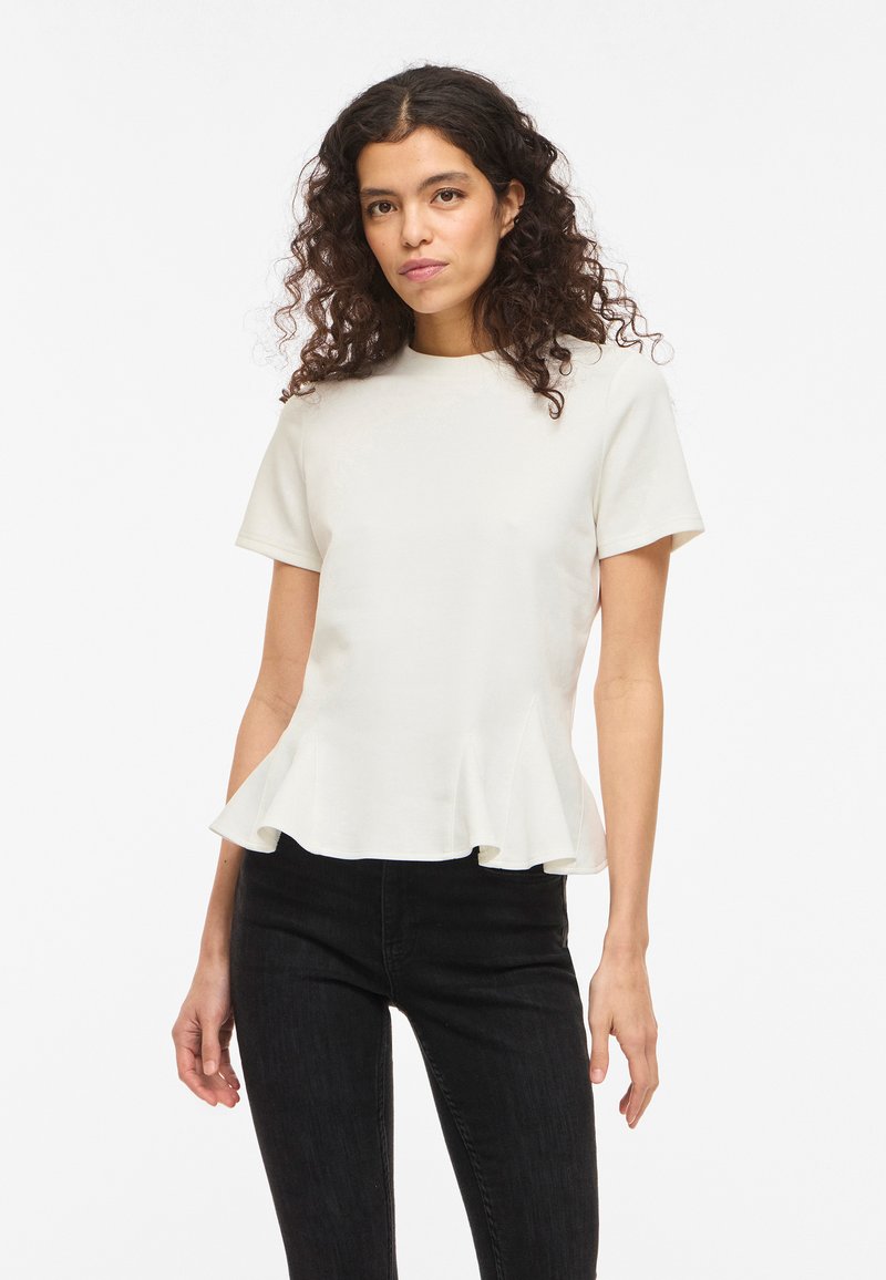 Kvinde med krøllet hår iført en hvid peplum-top med korte ærmer og sorte bukser, stående mod en ensfarvet hvid baggrund.