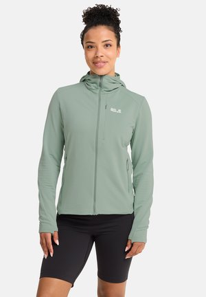 Lichtgroene jas met ritssluiting, verticale strepen, capuchon en zijzakken; gedragen over zwarte fietsshorts. Gladde, rekbare stof.