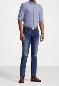 Camisa de cuadros azules y blancos con botones, vaqueros con las rodillas desgastadas, cinturón de cuero marrón y mocasines a juego.