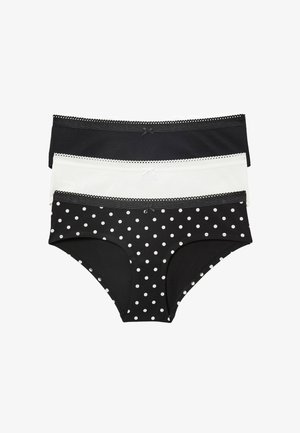 Femme portant un t-shirt blanc uni à manches courtes et un sous-vêtement noir à pois blancs sur un fond uni.